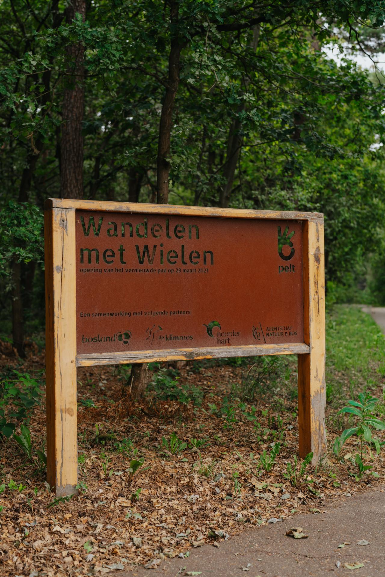 Houten buitenbord met gravure langs een wandelroute in het bos.